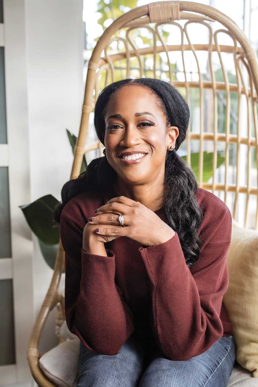  Ugochi smiling brightly in a maroon sweater with her elbows on her legs and her hands folded together.