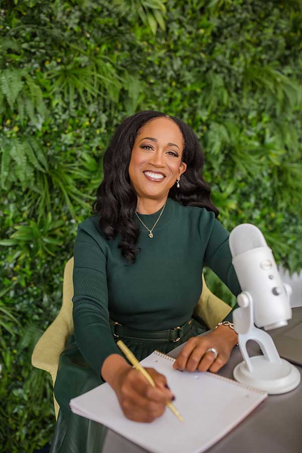 Ugochi in a green dress smiling in front of a podcast microphone, holding a pen with a book underneath