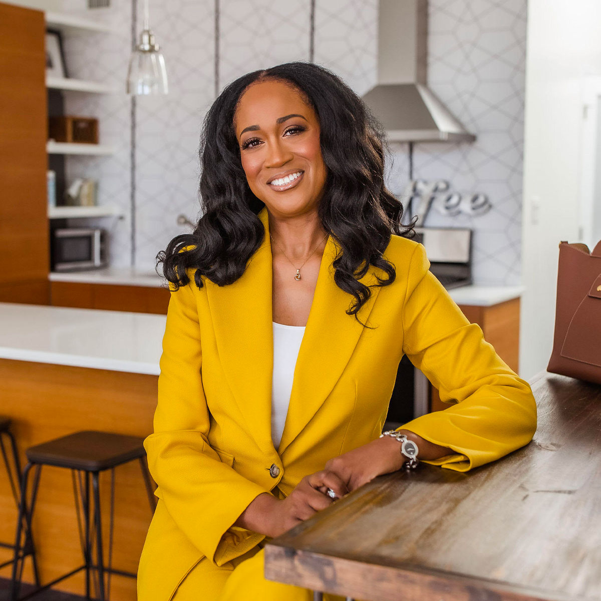 Ugochi sitting and smiling brightly, wearing a mustard color pantsuit.