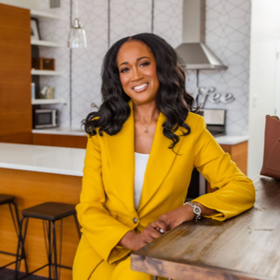 Ugochi sitting and smiling brightly, wearing a mustard color pantsuit.